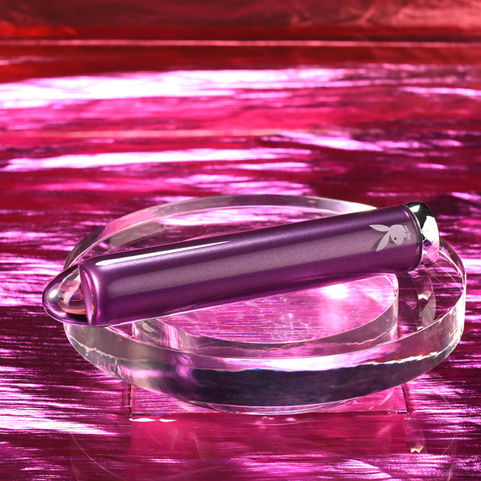 The Amethyst Slim Waterproof Rechargeable Glass Vibrator, featuring a small bunny logo, sits on a clear round platform against a shiny pink and magenta background.