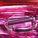 The Amethyst Slim Waterproof Rechargeable Glass Vibrator, featuring a small bunny logo, sits on a clear round platform against a shiny pink and magenta background.