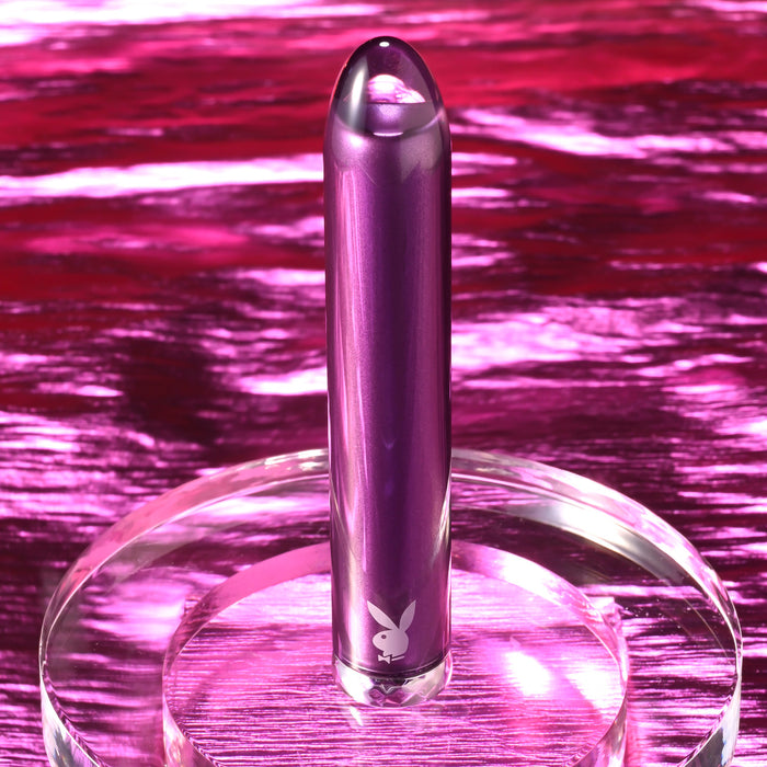 The Amethyst Slim Waterproof Rechargeable Glass Vibrator features a metallic purple finish with a Playboy bunny logo, displayed upright on a clear stand against a shiny pink backdrop—luxury and style for your collection.