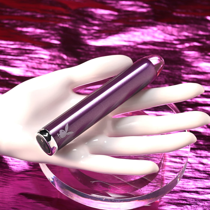 The Amethyst Slim Waterproof Rechargeable Glass Vibrator with a silver tip and Playboy bunny logo rests on a white mannequin hand against a pink metallic textured background.
