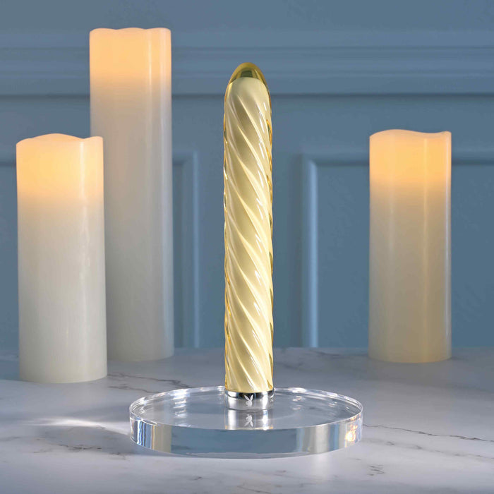 The Twist of Fate Slim Waterproof Rechargeable Glass Vibrator, featuring a textured spiral design and clear round base, stands upright beside three flameless candles on marble with a soft blue backdrop.