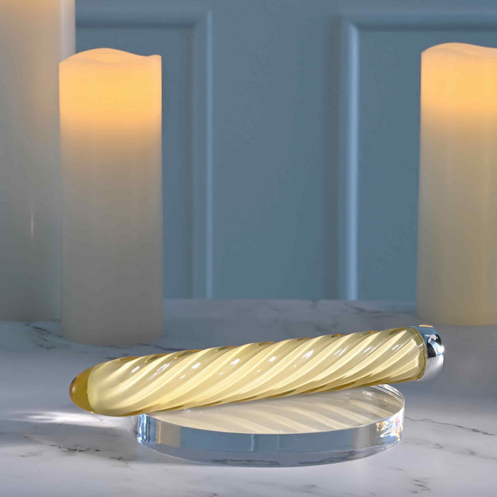 The Twist of Fate Slim Waterproof Rechargeable Glass Vibrator, featuring a cream-colored spiral glass design and metallic end, is shown on a clear round platform atop marble with three softly glowing pillar candles in the background.