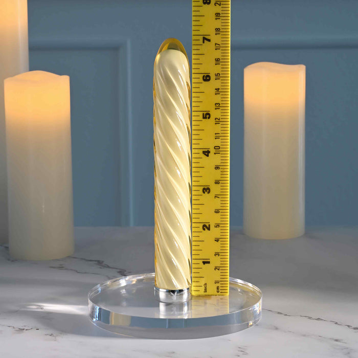 The Twist of Fate Slim Waterproof Rechargeable Glass Vibrator, cream-colored with a spiral glass design, stands upright on a clear round base beside a yellow measuring tape, with two glowing pillar candles on a marble surface.