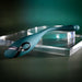The Pinpoint Perfection Flexible Slim Vibrator, a sleek teal device with a metallic accent and curved design, is displayed on a clear rectangular acrylic platform against a dark green background.