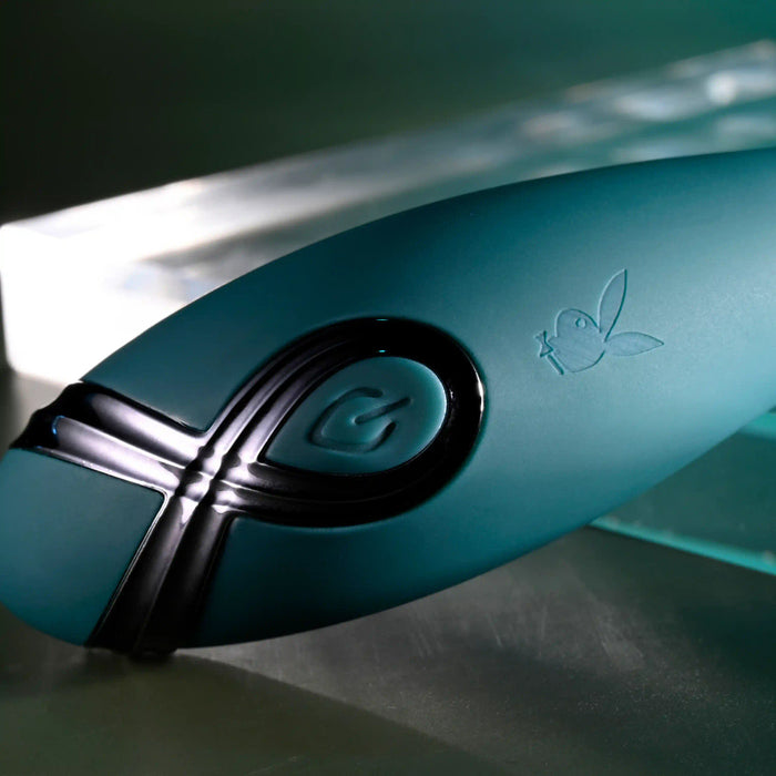 A close-up of the Pinpoint Perfection Flexible Slim Vibrator in teal shows its bendable, body-safe silicone surface with a power button and Playboy bunny logo, set against a blurred metallic background.