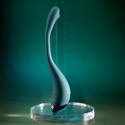 The Pinpoint Perfection Flexible Slim Vibrator, a sleek teal silicone device with a curved, smooth design, stands upright on a clear round base against a green gradient background.