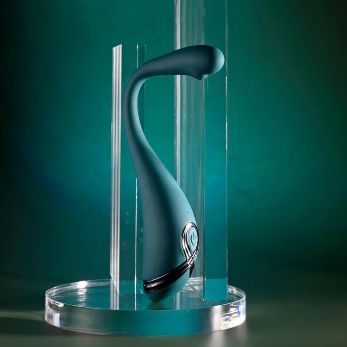 The Pinpoint Perfection Flexible Slim Vibrator, featuring a modern teal design with metallic accents and a curved, bendable shape, stands upright on a clear platform amid vertical acrylic panels against a teal backdrop.