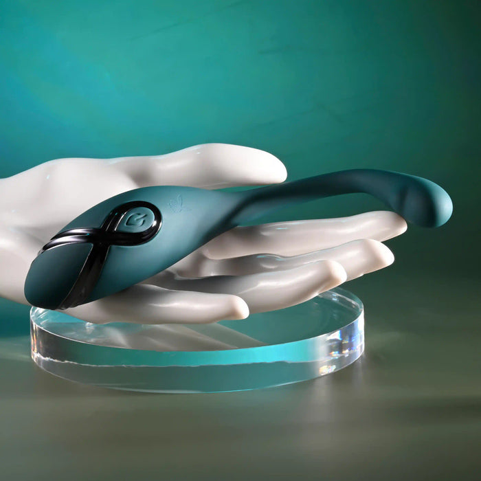 A white sculpted hand displays the Pinpoint Perfection Flexible Slim Vibrator—a sleek, teal, bendable toy made of body-safe silicone with a power button and black accent—set against a green gradient background.