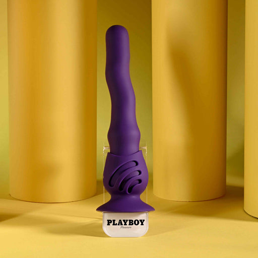 A purple Wiggle It Silicone Wiggling Vibrator with a suction cup base stands upright on a clear base labeled "PLAYBOY Pleasure," set against a yellow background with cylindrical yellow props.