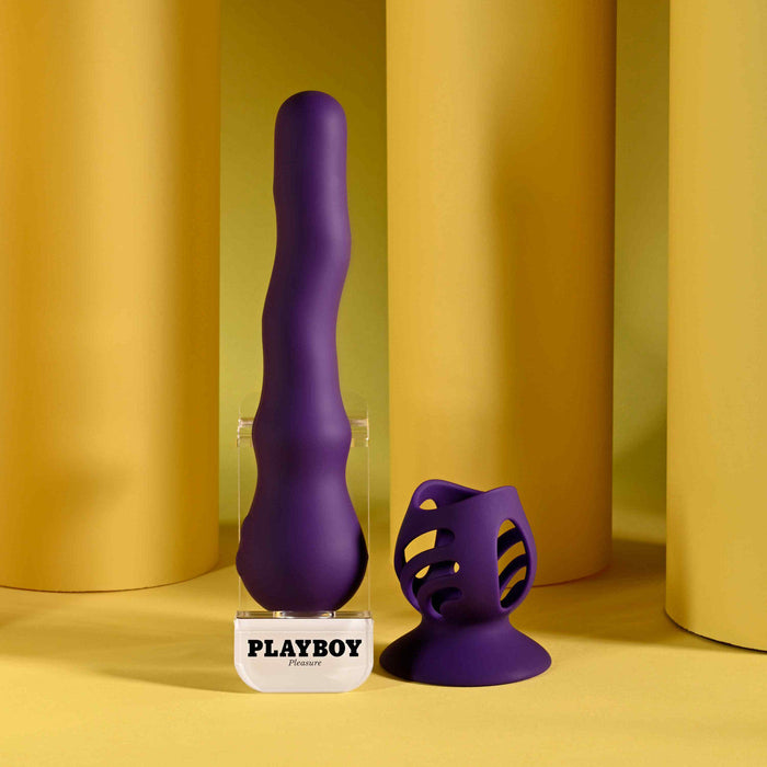 The Wiggle It Silicone Wiggling Vibrator with Suction Cup Base and matching accessory are shown on a yellow surface with cylindrical yellow props. The toy stands upright on its clear base labeled "PLAYBOY" in black letters.
