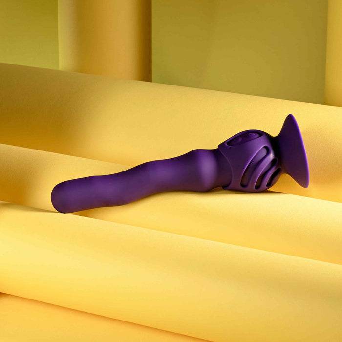 The Wiggle It Silicone Wiggling Vibrator with Suction Cup Base, featuring a purple, ergonomic design and textured grip, is displayed on rolled yellow paper against a yellow background.