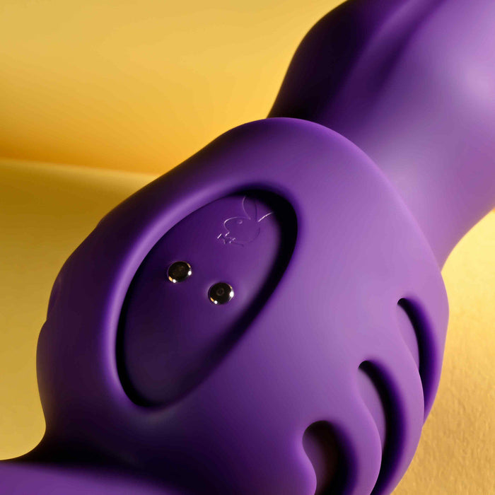 A close-up of the Wiggle It Silicone Wiggling Vibrator with Suction Cup Base, featuring a smooth, contoured purple design and two small metal charging ports, set against a yellow background.