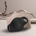 The Palm Vibrating Versatile & Accessible Masturbator, made of black body-safe silicone with a textured surface and dual openings, is displayed in front of a beige rock and dried wooden branches against a neutral background.