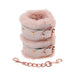 A pair of pink, fluffy Peaches 'n Cream Fur Handcuffs from Sportsheets, featuring rose gold buckles, are stacked together. Below is a short chain with clips on both ends that match the buckles. These bondage accessories are elegantly showcased on a plain white background.
