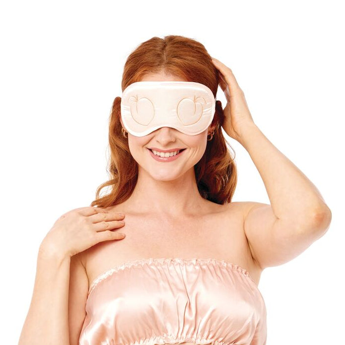 A smiling woman poses against a white background, wearing the Peaches 'n Cream Silky Blindfold with a light pink satin bandeau top and matching bottoms—making it the perfect date night accessory.