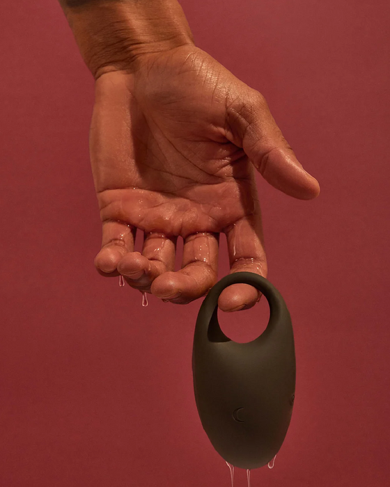A hand holding a black, egg-shaped, USB rechargeable Pepper Connect Vibrating Cock Ring made of body-safe silicone with a loop at the top, against a red background. Droplets of water are visible on the hand.