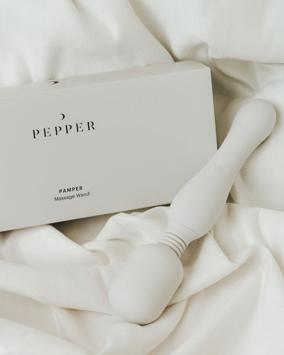 A sleek, USB rechargeable Pepper Pamper Massage Wand lies on soft, wrinkled fabric, next to its minimalist packaging labeled 'pepper'.