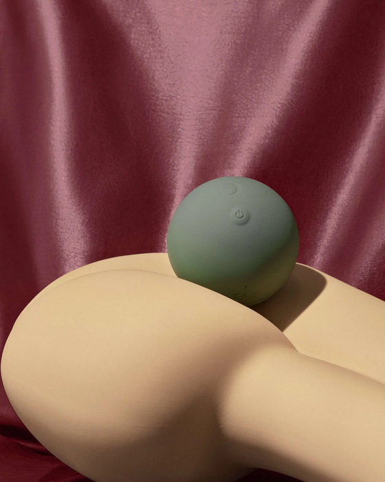 A modern still life composition featuring a Pepper Roll Vibrating Massager resting atop a curved, beige sculpture against a draped pink satin background, evoking a sense of minimalist elegance.