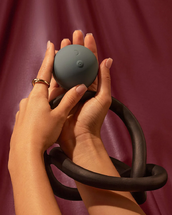 A pair of hands gently holding a Pepper Roll Vibrating Massager with a looped, flexible band attached, against a crimson draped background.