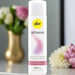 A bottle of Pjur Woman Concentrated Silicone Personal Lubricant 3.4 oz. sits on a table with blurred flowers in the background. The white bottle with yellow and pink accents is ideal for sensitive skin and easing vaginal dryness.