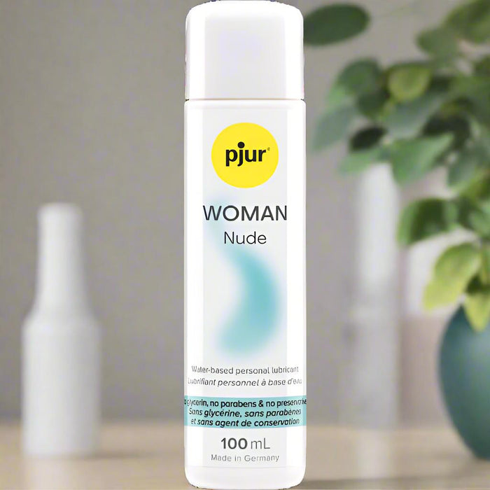 A 100 mL bottle of Pjur Woman Nude Water Based Lubricant is on a table, with a blurred green plant and white items behind it. The label notes it’s odor-free, ideal for sensitive skin, and made without parabens, glycerine, or preservatives.