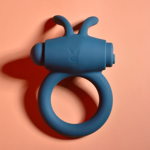 A blue Playboy Bunny Buzzer Vibrating Silicone Cock Ring, made from body-safe silicone with two short bunny ear-like protrusions and a subtle bunny logo, is shown on a peach-colored background.