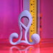 The Playboy Fantasy Cock Cage - Vibrating, Rechargeable, Silicone stands before a ruler on reflective surfaces with a pink and purple gradient backdrop, highlighting its unique shape under soft, colorful lighting.