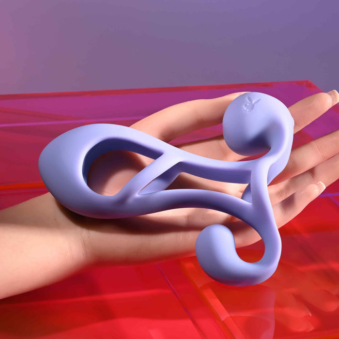 A hand holds the Playboy Fantasy Cock Cage—vibrating, rechargeable, and made of silicone—with modern curved and looped shapes against a pink and purple background.