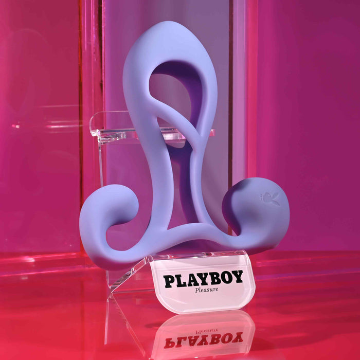 The Playboy Fantasy Cock Cage—vibrating, rechargeable, and made of silicone—is shown on a clear stand labeled "PLAYBOY Pleasure" against a vibrant pink background.