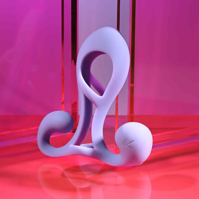 The Playboy Fantasy Cock Cage, a sculptural lavender vibrating silicone toy, stands upright on a glossy pink and red surface against a vivid pink and purple background.