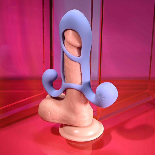 The Playboy Fantasy Cock Cage - Vibrating, Rechargeable, Silicone is shown upright on a vivid pink and orange background, featuring a blue silicone cage with rounded curves wrapping and extending from the shaft for playful couple’s stimulation.