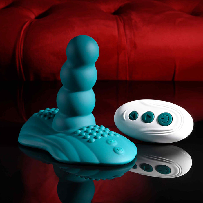 The Playboy Have a Seat Remote Control Grinding Vibrator Pad, made from body-safe silicone with a beaded shaft and textured suction cup base, is displayed next to its white remote on a glossy black surface against a red velvet backdrop.