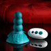 The Playboy Have a Seat Remote Control Grinding Vibrator Pad with its sleek design sits on a glossy black surface next to its white remote, set against a striking red tufted background.