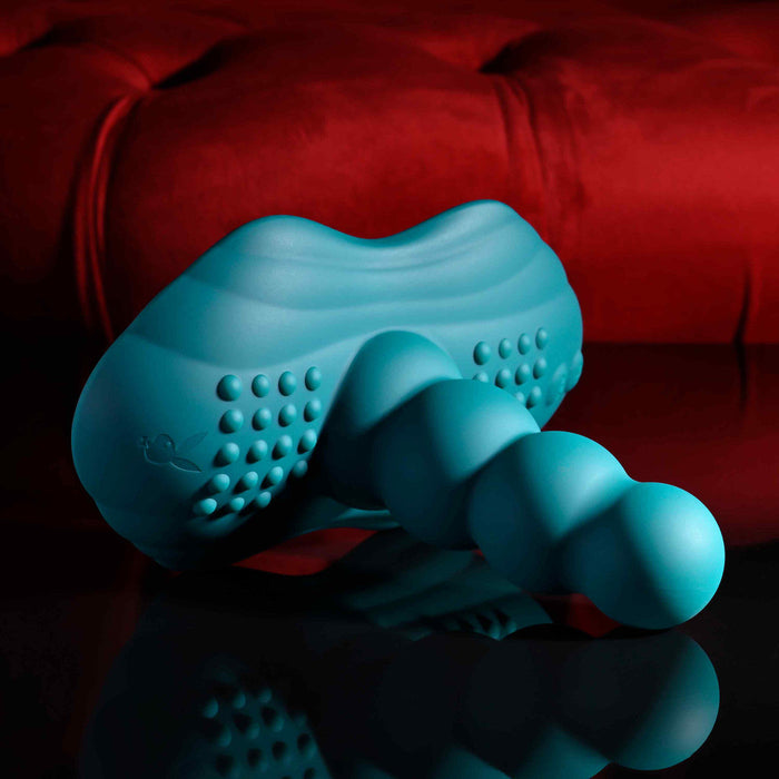The Playboy Have a Seat Remote Control Grinding Vibrator Pad, crafted from body-safe silicone with textured nubs and a turquoise beaded design, rests on a reflective black surface before a plush red backdrop, featuring hands-free suction cup versatility.