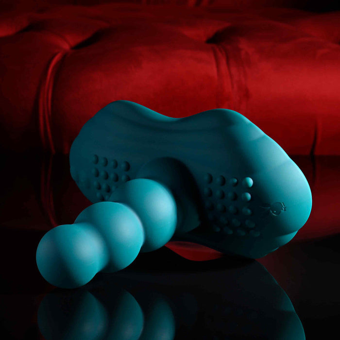 The Playboy Have a Seat Remote Control Grinding Vibrator Pad, a teal silicone personal massager with ribbed texture, rests on a glossy black surface before a red tufted background.