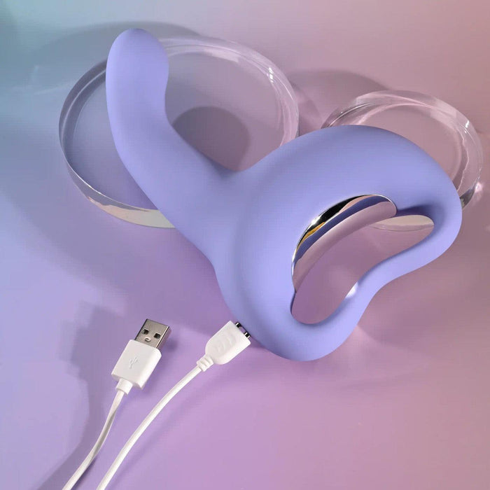 The Playboy Nice to Meet You Powerful Thrusting Vibrator with Handle, shown in lavender silicone with a curved design, is displayed on a pastel background. It is connected to a white USB charging cable, with two clear round objects nearby.