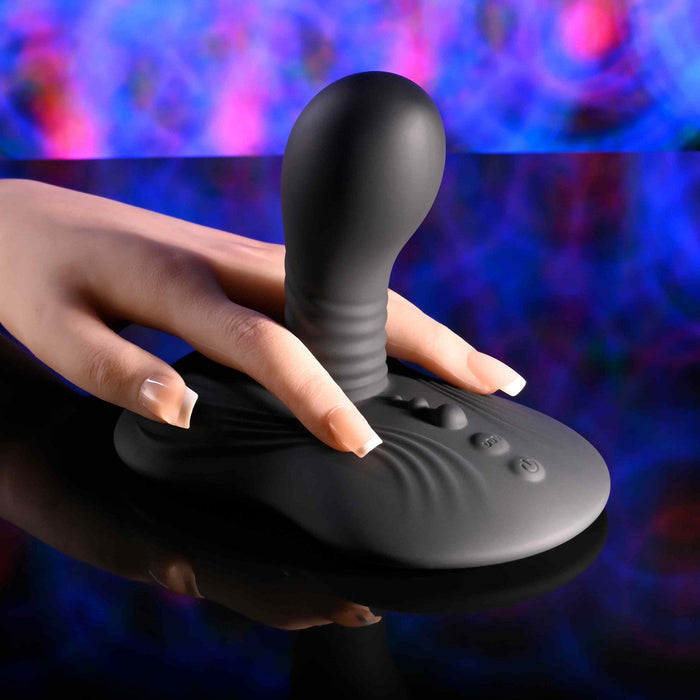 A hand with manicured nails touches the Playboy Sittin' & Spinnin' Hands Free G-Spot Vibrating Grinder Pad, a black silicone toy with a bulbous top, resting on a glossy surface against a blue and pink blurred background.