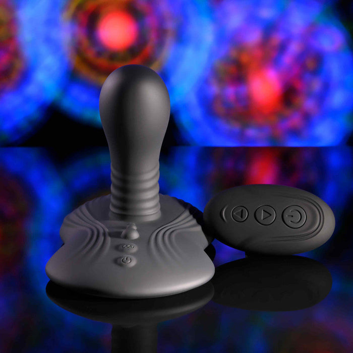 The Playboy Sittin' & Spinnin' Hands Free G-Spot Vibrating Grinder Pad and its remote control rest on a reflective surface, illuminated by blurred blue, red, and purple lights in the background.