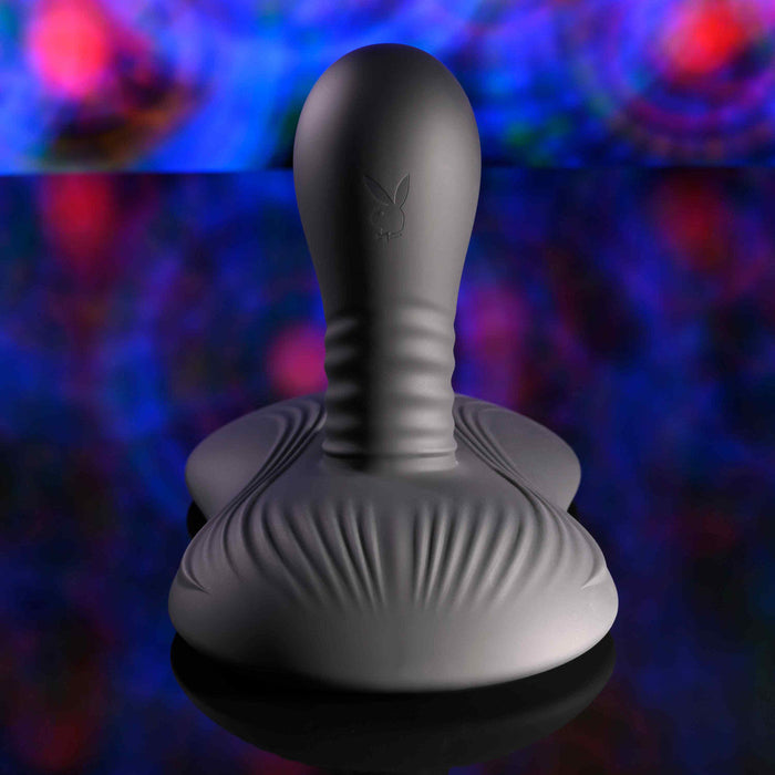 The Playboy Sittin' & Spinnin' Hands Free G-Spot Vibrating Grinder Pad, featuring a gray sculpted design with a rounded top, textured surface, and the iconic bunny logo, sits on a reflective surface against an abstract multicolored background.