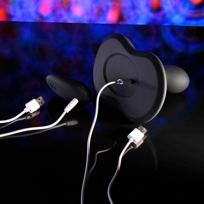 The Playboy Sittin' & Spinnin' Hands Free G-Spot Vibrating Grinder Pad, a sleek black pleasure device with rounded edges, is connected to a white USB charging cable and displayed on a reflective surface with vibrant blue and red lights in the background.