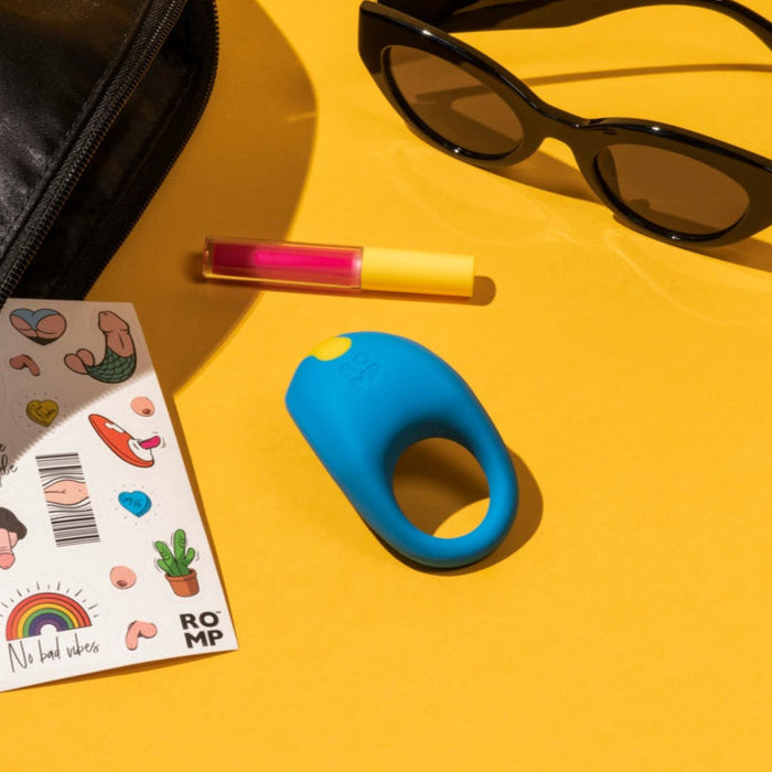 A Romp Juke Silicone Vibrating Couples Cock Ring, black sunglasses, pink lip gloss, and colorful stickers are arranged on a yellow surface near a black bag.