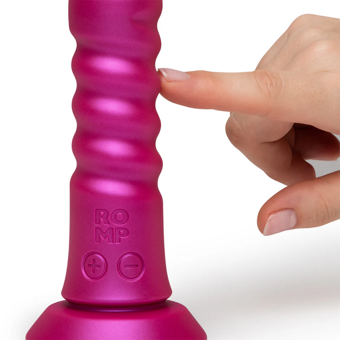 A hand with light skin gently touches the ROMP Jiggle Thrusting Vibrator with Suction Base, which stands upright, features "+" and "–" buttons, ribbed pink design, and "ROMP" embossed near the base.