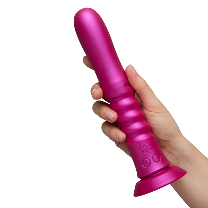 A hand holds the ROMP Jiggle Thrusting Vibrator with Suction Base—a bright pink, ribbed silicone toy featuring hands-free pleasure and the "ROMP" logo near the base.