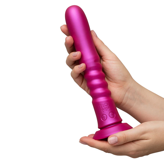 Two hands hold the ROMP Jiggle Thrusting Vibrator with Suction Base, a large metallic pink ribbed silicone toy, vertically positioned against a plain white background.