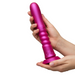Two hands hold the ROMP Jiggle Thrusting Vibrator with Suction Base, a large metallic pink ribbed silicone toy, vertically positioned against a plain white background.