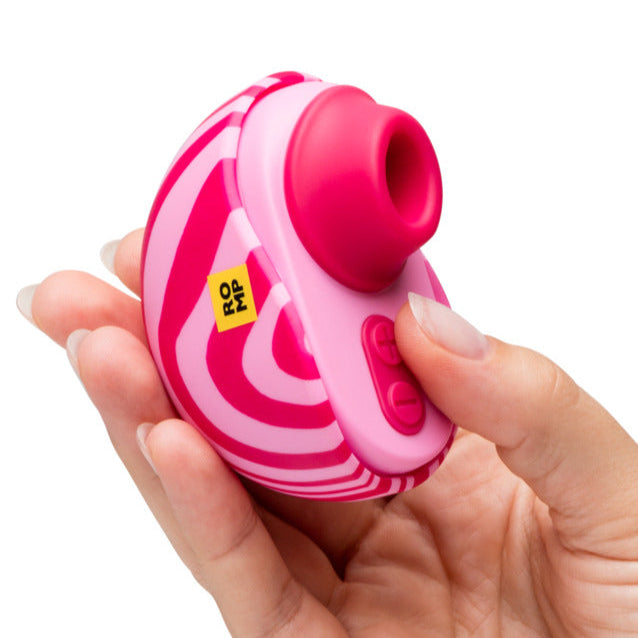 A hand holds the Romp Spin Small Discreet Pleasure Air Clitoral Stimulator, a pink and white striped, oval travel-size device with buttons, a circular opening, and a yellow "ROMP" label. It uses unique Pleasure Air technology for enhanced satisfaction.