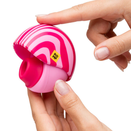 A person holds the Romp Spin Small Discreet Pleasure Air Clitoral Stimulator, a round, pink and red striped travel-size device, in one hand while pressing the button on top with the other.
