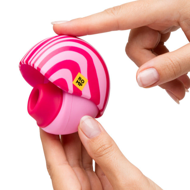 A person holds the Romp Spin Small Discreet Pleasure Air Clitoral Stimulator, a round, pink and red striped travel-size device, in one hand while pressing the button on top with the other.