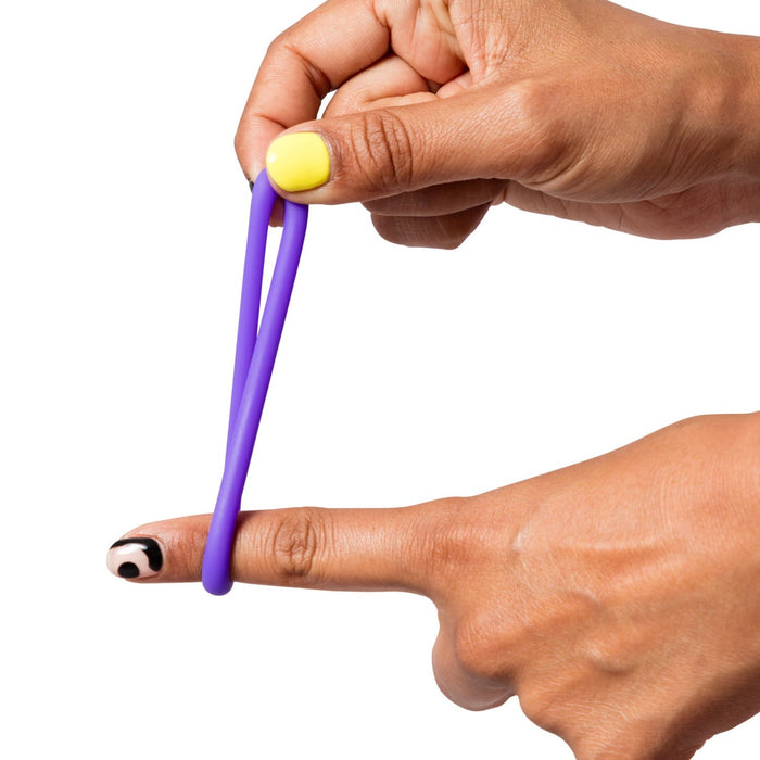 A hand with yellow-painted nails stretches a purple Romp Remix Cock Ring Trio over the finger of another hand, which has a playful eye nail design, against a white background.