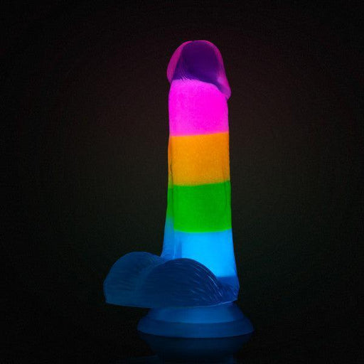 The Rainbow Rave Slim Glow in the Dark 6.5 Inch Silicone Dildo features a realistic shape, vibrant glow-in-the-dark rainbow stripes, is made from silicone, and is strap-on harness compatible for versatile play.